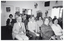 B&W photo of a small group of people sitting on rows of chairs in a room. Two women in the second row are looking directly at the camera. In the background, there is a tall filing cabinet in the corner, near a large window. On the other side of the window, there is a large multipanel display board. There are a few pictures and/or posters and a large framed mirror on the walls.