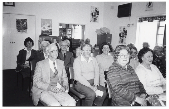 B&W photo of a small group of people sitting on rows of chairs in a room. Two women in the second row are looking directly at the camera. In the background, there is a tall filing cabinet in the corner, near a large window. On the other side of the window, there is a large multipanel display board. There are a few pictures and/or posters and a large framed mirror on the walls.