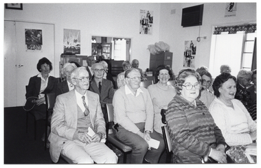 B&W photo of a small group of people sitting on rows of chairs in a room. Two women in the second row are looking directly at the camera. In the background, there is a tall filing cabinet in the corner, near a large window. On the other side of the window, there is a large multipanel display board. There are a few pictures and/or posters and a large framed mirror on the walls.