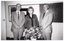 B&W photo of three people, standing next to one another, posing for a photo. There is a large floral bouquet on a small table in front of them. On the wall behind them, there is a large display board and blackboard. There is an open door (partially obscured) to the left of the frame.