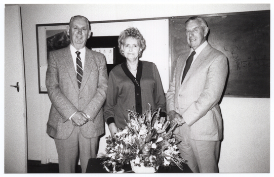 B&W photo of three people, standing next to one another, posing for a photo. There is a large floral bouquet on a small table in front of them. On the wall behind them, there is a large display board and blackboard. There is an open door (partially obscured) to the left of the frame.
