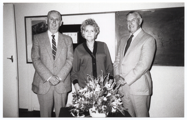 B&W photo of three people, standing next to one another, posing for a photo. There is a large floral bouquet on a small table in front of them. On the wall behind them, there is a large display board and blackboard. There is an open door (partially obscured) to the left of the frame.