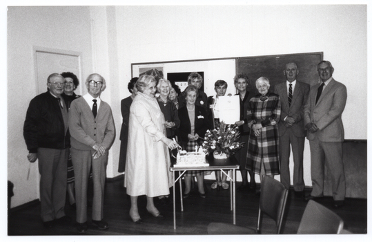 B&W wide frame photo of a group of people posing for a photo around a large rectangular cake and a floral bouquet. The cake and bouquet are on a small card table. One woman is holding a knife which has been plunged into the cake. Another woman is holding up a large certificate. On the wall behind them, there is a large display board and blackboard. There is a closed door (partially obscured) to the left of the frame. There are some chairs in the foreground.