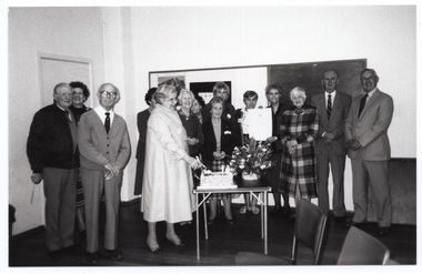 B&W wide frame photo of a group of people posing for a photo around a large rectangular cake and a floral bouquet. The cake and bouquet are on a small card table. One woman is holding a knife which has been plunged into the cake. Another woman is holding up a large certificate. On the wall behind them, there is a large display board and blackboard. There is a closed door (partially obscured) to the left of the frame. There are some chairs in the foreground.
