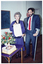 COL photo of two people posing for a photo beside a floral bouquet and a rectangular cake on a card table. The woman is holding a large unframed certificate and the man is leaning on one crutch. There is a third person standing behind them. In the background, there is a blackboard on the wall and a closed door further along the wall (both partially obscured).