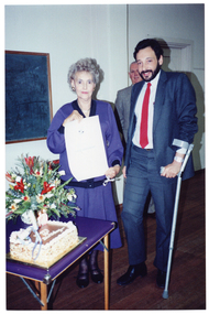 COL photo of two people posing for a photo beside a floral bouquet and a rectangular cake on a card table. The woman is holding a large unframed certificate and the man is leaning on one crutch. There is a third person standing behind them. In the background, there is a blackboard on the wall and a closed door further along the wall (both partially obscured).