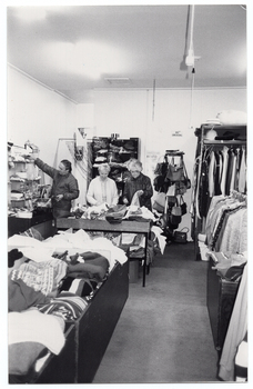 B&W photo of three people inside a shop. In the foreground, there are several large display bins filled with clothing in the centre aisle of the store, while shelving filled with miscellaneous goods have been placed against the walls. At the rear of the store, two women are sorting and tidying clothes from one of the clothing display bins while, beside them, a man is perusing items from one of the shelves.