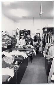 B&W photo of three people inside a shop. In the foreground, there are several large display bins filled with clothing in the centre aisle of the store, while shelving filled with miscellaneous goods have been placed against the walls. At the rear of the store, two women are sorting and tidying clothes from one of the clothing display bins while, beside them, a man is perusing items from one of the shelves.