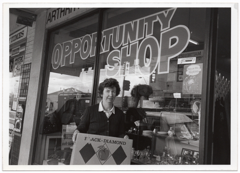 B&W photo of a woman standing in front of a shop window. She is holding a cardboard box printed with the text "BLACK DIAMOND" and a drawing of an apple between two black diamond shapes. Behind her, the text "ARTHR..." (partially obscured) is visible on the exterior wall above the front window. On the window is the text "OPPORTUNITY SHOP". Inside the front window are various displays, including crockery, glassware, and bric-a-brac.
