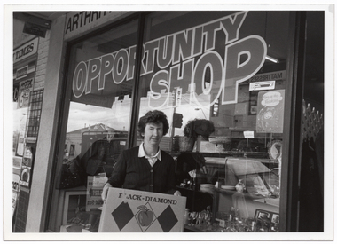 B&W photo of a woman standing in front of a shop window. She is holding a cardboard box printed with the text "BLACK DIAMOND" and a drawing of an apple between two black diamond shapes. Behind her, the text "ARTHR..." (partially obscured) is visible on the exterior wall above the front window. On the window is the text "OPPORTUNITY SHOP". Inside the front window are various displays, including crockery, glassware, and bric-a-brac.