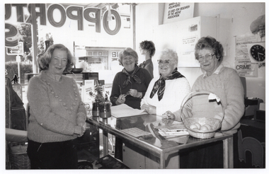 B&W photo of four women inside a shop. One woman is standing in front of the counter (a glass display cabinet), while the other three women are standing behind it. They are all looking directly at the camera and smiling. On top of the glass counter, there are some brochures on a stand, a sales book, some printed booklets stacked loosely, and some fresh fruit in a cane basket. One of the brochures is titled, "FIGHTING ARTHRITIS". Behind the women, is the front shop window, with the painted text, "OPPORTUNITY SHOP" (partially obscured), appearing in reverse. On the wall behind them, there is a small sign resting against the wall above the meter box. The text (partially obscured) reads, "...WHICH IS... VOLUNTEERS... IS USED FOR RESEARCH BY THE ABOVE ASSOCIATION."