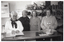 B&W photo of four women inside a shop. They are standing behind a glass display cabinet, which serves as the sales counter. They are all facing the camera and smiling. On the wall behind them are shelves of soft toys and books. On a meter box affixed to the wall is a handwritten sign which says, "Please do not ask for price reduction. This is a charity shop staffed by volunteers. A.F.V." and another sign which says "NO EXCHANGE".