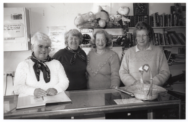 B&W photo of four women inside a shop. They are standing behind a glass display cabinet, which serves as the sales counter. They are all facing the camera and smiling. On the wall behind them are shelves of soft toys and books. On a meter box affixed to the wall is a handwritten sign which says, "Please do not ask for price reduction. This is a charity shop staffed by volunteers. A.F.V." and another sign which says "NO EXCHANGE".