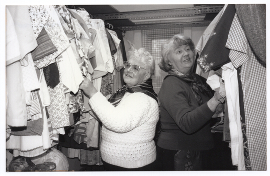 B&W photo of two women standing between two hanging clothes racks full of clothes. They are standing close, with their backs to one another, perusing the garments at close range.