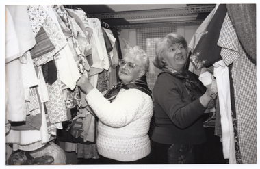 B&W photo of two women standing between two hanging clothes racks full of clothes. They are standing close, with their backs to one another, perusing the garments at close range.