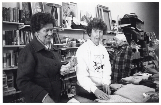 B&W photo of three women standing inside a shop. Two of the women are standing behind a counter with folded woollen jumpers displayed on the top. They are both facing the camera, but one of them is looking down at a small doll that she is holding. The third woman is in the background, facing a wall of shelving filled with books, bric-a-brac, crockery, glassware, framed prints and empty picture frames, and other miscellaneous items. Further back along the wall, there is a rack of handbags and shoulder bags.