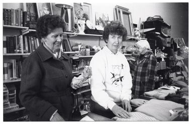 B&W photo of three women standing inside a shop. Two of the women are standing behind a counter with folded woollen jumpers displayed on the top. They are both facing the camera, but one of them is looking down at a small doll that she is holding. The third woman is in the background, facing a wall of shelving filled with books, bric-a-brac, crockery, glassware, framed prints and empty picture frames, and other miscellaneous items. Further back along the wall, there is a rack of handbags and shoulder bags.