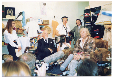 COL photo of a elderly man, dressed in an old brown checked dressing gown, sitting on a art deco club lounge armchair in a crowded room. He is surrounded by several people, including two television camera operators and a boom microphone operator. Sitting near him is an unidentified man with grey hair and beard, wearing a navy blue three-piece suit. A woman wearing a white shirt and navy blue skirt is standing nearby. In the background, shelves filled with second hand items and cardboard boxes line the walls. In the foreground, several people's heads (Photographed from behind) are evident.
