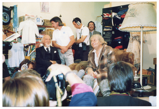 COL photo of a elderly man, dressed in an old brown checked dressing gown, sitting on a art deco club lounge armchair in a crowded room. He is surrounded by several people, including two television camera operators and a boom microphone operator. Sitting near him is an unidentified man with grey hair and beard, wearing a navy blue three-piece suit. A woman wearing a white shirt and navy blue skirt is standing nearby. In the background, shelves filled with second hand items and cardboard boxes line the walls. In the foreground, several people's heads (Photographed from behind) are evident.