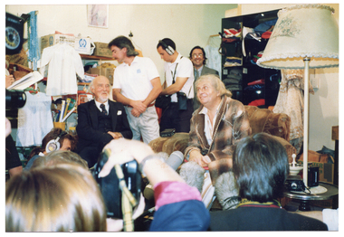 COL photo of a elderly man, dressed in an old brown checked dressing gown, sitting on a art deco club lounge armchair in a crowded room. He is surrounded by several people, including two television camera operators and a boom microphone operator. Sitting near him is an unidentified man with grey hair and beard, wearing a navy blue three-piece suit. A woman wearing a white shirt and navy blue skirt is standing nearby. In the background, shelves filled with second hand items and cardboard boxes line the walls. In the foreground, several people's heads (Photographed from behind) are evident.
