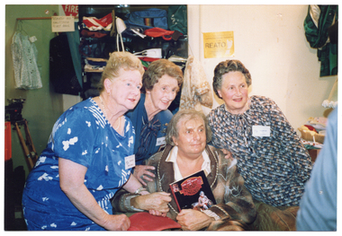 COL photo of a elderly man, dressed in an old brown checked dressing gown, sitting on a art deco club lounge armchair in a crowded room. He is surrounded by three middle-aged women, each wearing a name tag that says "Volunteer". He is holding the hand of one of the women with one hand, and a book with his other hand. The book is titled, "The Life and Death of Sandy Stone". In the background, there are miscellaneuous items protruding from a large hanging wall pocket storage organiser, a shirt handing from a rod in the corner, and other miscellaneous items around the room.