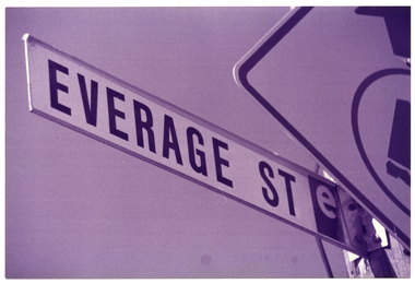 B&W photo of a street sign name "Everage St". Another road sign, for a pedestrian crossing (partially obscured) is also in the frame. The photo has been taken from a low angle.