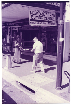 B&W photo of a woman and a man at a suburban shopping strip. They are walking along the footpath towards one another. She is dressed in a long evening gown and is carrying a bunch of long-stemmed flowers, the top of which are resting over one shoulder. He is dressed in a shirt and trousers, and is looking down at the single long-stem of flowers that he is carrying. Hanging from the overhead veranda ceiling above are a sign and a banner. The sign (partially obscured) says, "...H CONVERTERS".  The banner says, "CA$H CONVERTERS NEW DRIVE THRU BUYING CENTRE REAR LANE EASY ACCESS". There is a narrow laneway (partially obscured) beside the building.