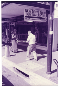 B&W photo of a woman and a man at a suburban shopping strip. They are walking along the footpath towards one another. She is dressed in a long evening gown and is carrying a bunch of long-stemmed flowers, the top of which are resting over one shoulder. He is dressed in a shirt and trousers, and is looking down at the single long-stem of flowers that he is carrying. Hanging from the overhead veranda ceiling above are a sign and a banner. The sign (partially obscured) says, "...H CONVERTERS".  The banner says, "CA$H CONVERTERS NEW DRIVE THRU BUYING CENTRE REAR LANE EASY ACCESS". There is a narrow laneway (partially obscured) beside the building.