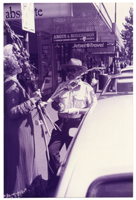 B&W photo of a woman and a man at a suburban shopping strip. She is wearing a long evening gown and is carrying a bunch of long-stemmed flowers. He is wearing a parking inspector's uniform and is holding a pen poised over a pad of paper. Several shop signs are hanging from the verandah ceiling above them, including "absolute", "ANGUS & ROBERTSON", and "Jetset Travel". In the distance, there are several people walking along the footpath.