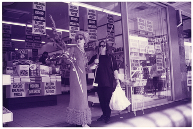 B&W photo of two woman at a suburban shopping strip. One woman is wearing a long evening gown, a long beaded necklace and novelty sunglasses in the shape of two swans. She is carrying a bunch of long-stemmed gladioli flowers. The other woman is dressed in dark coloured t-shirt, trousers, and shoes. They are standing in front of a bookshop. There are numerous posters throughout the shop and in the shop window with the text, "RECORD BREAKING PRICES RECORD BREAKERS RECORD BREAKING SAVINGS".