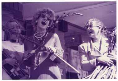 B&W medium close up photo of three women at a suburban shopping strip. The woman in the centre is wearing a long evening gown, a long beaded necklace and novelty sunglasses in the shape of two swans. She is carrying two long-stems of gladioli flowers, which is holding crossed in an x-formation across her chest. The other two women, standing on either side of her, are dressed in contemporary clothes - one is wearing a jacket. They are both wearing novelty glasses in the shape of two birds. They are each carrying a large bunch of long-stemmed gladioli flowers. All three women are laughing.