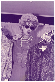 B&W photo of a woman holding up some clothes on hangers inside a shop. She is wearing a long evening gown, a long beaded necklace and novelty sunglasses in the shape of two swans. Behind her are some racks of clothes.