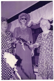 B&W photo of three women inside a shop. The woman in the centre is dressed in a long evening gown, a long beaded necklace and novelty sunglasses in the shape of two swans. She is flanked by two other women who are dressed in contemporary day dresses in printed fabrics. The two women are each holding a flowering gladioli stem. In the background, there is a lace-edge valance hanging across the ceiling.