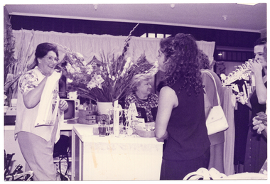 B&W photo of several women inside a shop. One woman, who is standing behind a counter, is handing over a plastic bag to another woman. Behind them, there is another woman either sitting down or leaning on the other side of the counter. To the right of the frame, there are three other women standing near the counter. One of them (partially obscurred) appears to be talking to the woman who is sitting/leaning on the counter. The other two women (one partially obscurred) appear to be waiting to be served. There is a large bouquet of flowering gladioli stems in a large vase on the counter.