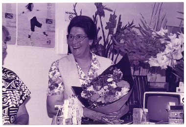 B&W medium close-up photo of a woman holding a bouquet of flowers. Beside her stands another woman (partially obscured). On the counter in front of her is a large bouquet of flowering gladioli stems in a vase, a small portable television set, and various other items. One of these is a small box or container, on which there is some handwritten text. The heading of the text is "ARTHRITIS OP SHOPS!!", beneath which there is the Arthritis Foundation of Victoria logo and some more text. On the wall behind them, there are some posters and a 'No Smoking' sign.