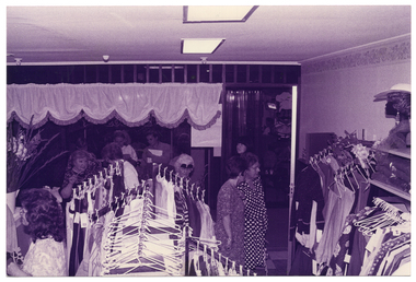 B&W photo of several people inside a shop. There are multiple racks of clothes hanging on coathangers. To the right of the frame, there is some shelving against the wall containing some folded clothing and a display mannequin head showcasing a hat and wig. To the left of the frame, there is a large bouquet of gladioli flowers in a large vase on a countertop. In the background, there is a lace-edged valance across the top of a wooden rail hanging from the ceiling. The photo has been taken from a high angle, above floor level.