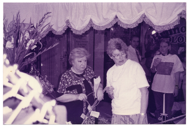 B&W photo of four people inside and outside some shops. In the foreground, two women are in one shop, looking down towards some items. To the left of the women, there is a large gladioli flower arrangement (partially obscured). In the background, to the right of the frame, a man and a woman are standing nearby. They appear to be outside the shop window, looking in. There is another shopfront window evident behind them. The woman is holding a sandwich or burger in one hand.