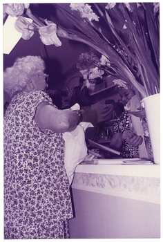 B&W photo of several people inside a shop. In the foreground, one woman is standing behind a counter. She is holding a plastic bag. On the other side of the counter, there are two women standing (partially obscured). Another person, with their back to the camera, is reaching for something on a shelf along the wall. In the foreground, to the left of the frame, there is a large bouquet of flowering gladioli stems in a vase on the countertop. The photo has been taken from behind the counter, at a low angle.