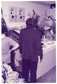 B&W photo of two people looking at a framed painting or print, which someone (partially obscured) is holding up. The person holding the picture is standing behind a counter. There are various items on the countertop, such as a small portable television set, a book, a miniature set of drawers, a large arrangement of long-stemmed gladioli flowers, three glasses of wine, and a brochure. In the foreground, to the left of the frame, there are several small baskets of miscellaneous items on top of a table or countertop (obscured). There are two identical posters on the wall behind the counter. The title of the posters is, "Step out for strong bones".