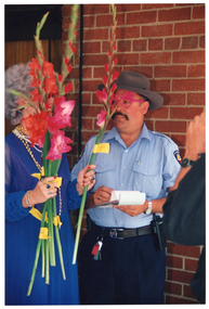 COL photo of a man and a woman. The woman is wearing a royal blue evening dress, with two beaded necklaces around her neck. She is holding several long-stemmed gladioli flowers in one hand as she hands one to the man. He is wearing a traffic inspector's uniform, with some bright pink-framed glasses. He is holding a pen, which is poised over a pad of paper. In the foreground, to the right of the frame, a man wearing a black shirt (partially obscured) is holding one hand up near his face.