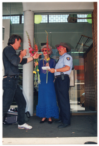 COL photo of three people standing in an alcove outside some shops. In the centre is a woman. She is wearing a royal blue evening dress, with two beaded necklaces around her neck and a pair of red novelty glasses in the shape of two swans. She is holding several long-stemmed gladioli flowers in one hand, and a single gladioli stem in her other hand. To the right of the frame, there is a man wearing a traffic inspector's uniform with some bright pink novelty glasses. He is holding a pen, which is poised over a pad of paper. To the left of the frame, there is a man wearing a black shirt and black trousers. He is holding a camera in one hand, and pointing to the other two people with his other hand.