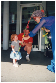 COL photo of a woman handing a long flowering gladioli stem to a small child, while a man looks on. They are on the footpath outside a shop or business premises. The woman is wearing a royal blue evening dress, a wig, beaded necklaces, and red novelty glasses. She is bending forward towards the child. The child is wearing a grey and white striped t-shirt and a denim skirt. She is looking up at the woman as she grasps the long flower stem. The man is wearing a red t-shirt, black shorts, and runners. He is crouching down beside the child.