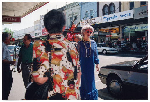 COL photo of several people at a suburban shopping strip. To the right of the frame is a woman. She is wearing a royal blue evening dress, a wig, beaded necklaces, and red novelty glasses. There is someone walking behind her, in the opposite direction. To the left of the frame, there are two men are talking to one another. In the foreground, in the centre of the frame, a person with a black shoulderbag on their shoulder is walking past. Behind her, another person (completely obscured) is carrying a bunch of long flowering gladioli stems. There are a few men standing or walking past. One of them is holding a flowering gladioli stem. In the background, several retail shops (such as Ferguson Plarre Bakehouses and Speeds Shoes) are evident across the street.