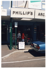 COL photo of the entrance to a shopping arcade in a suburban shopping strip. A sign above the arcade entrance says, "PHILLIP'S ARCADE" (partially obscured). At the entrance to the arcade, there is an A-frame sandwich board which contains the text, "EXPERT JEWELLERY SOLDERS WATCH BATTERIES AVAILABLE". On the footpath outside of the arcade entrance, there is another A-frame sandwich board. It contains the text, "Move it or Lose it", followed by a logo. Two women are walking across the arcade entrance in opposite directions; one of them is pushing a pram with a young child in it.