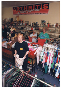 COL photo of four women inside a shop. They are sorting through clothing and other items. In the foreground, one woman is holding a small toddler's dress on a coathanger. She is looking up at the camera. There are racks of hanging clothes in front of and behind her. In the backgound, two women are holding up a small red t-shirt and a red bag, respectively. They are standing behind a row of large square display bins. Another woman is standing further back in the same aisle. On the wall behind the three women are rows of shelving filled with glassware, crockery, bric-a-brac, framed pictures, and various other miscellaneous items. On the wall above the shelving, there is a large orange, black, and white sign just below the ceiling. The text on the sign says, "ARTHRITIS", "Something can be done!" [the word 'can' is underlined], "ARTHRITIS FOUNDATION OF VICTORIA PHONE 862 2555".