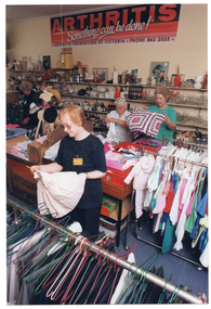 COL photo of four women inside a shop. They are sorting through clothing and other items. In the foreground, one woman is holding a small toddler's dress on a coathanger. She is looking down at the dress. There are racks of hanging clothes in front of and behind her. In the backgound, two women are holding up a crocheted blanket and a small item of clothing, respectively. They are standing behind a row of large square display bins. Another woman is standing further back in the same aisle. She is turning away from the camera. On the wall behind the three women, there is a large orange, black, and white sign just below the ceiling. The text on the sign says, "ARTHRITIS", "Something can be done!" [the word 'can' is underlined], "ARTHRITIS FOUNDATION OF VICTORIA PHONE 862 2555".