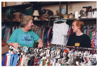 COL photo of two women standing between several racks of hanging clothes inside a shop. The woman on the left is wearing a jade green Club Med t-shirt, and an aqua name badge with the text, "VOLUNTEER Arthritis Foundation of Victoria". The woman on the right is wearing a plain black t-shirt, and a yellow name badge with the text, "SUPERVISOR Arthritis Foundation of Victoria". Above the clothing rack behind them, there are several shelves on the wall containing basketry, electrical kitchen appliances, a lamp shade, and other miscellaneous items.