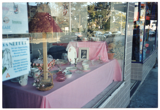 COL photo of a window display in the front window of a shop. The display features a pink floral on white/cream dinner set, some white crockery, and some glassware. The photo has been taken from outside the shop, so a partial image of the street scape is reflected on the glass window. There is another front display window in the background. The two display windows are surrounded by small grey square tiles, and there is a doorway in between.