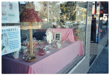 COL photo of a window display in the front window of a shop. The display features a pink floral on white/cream dinner set, some white crockery, and some glassware. The photo has been taken from outside the shop, so a partial image of the street scape is reflected on the glass window. There is another front display window in the background. The two display windows are surrounded by small grey square tiles, and there is a doorway in between.