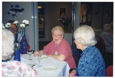 COL photo of a group of people sitting around a table. There are wine glasses, a vase of flowers, and small plates on the table. In the background, there are several posters and small pictures on the walls.